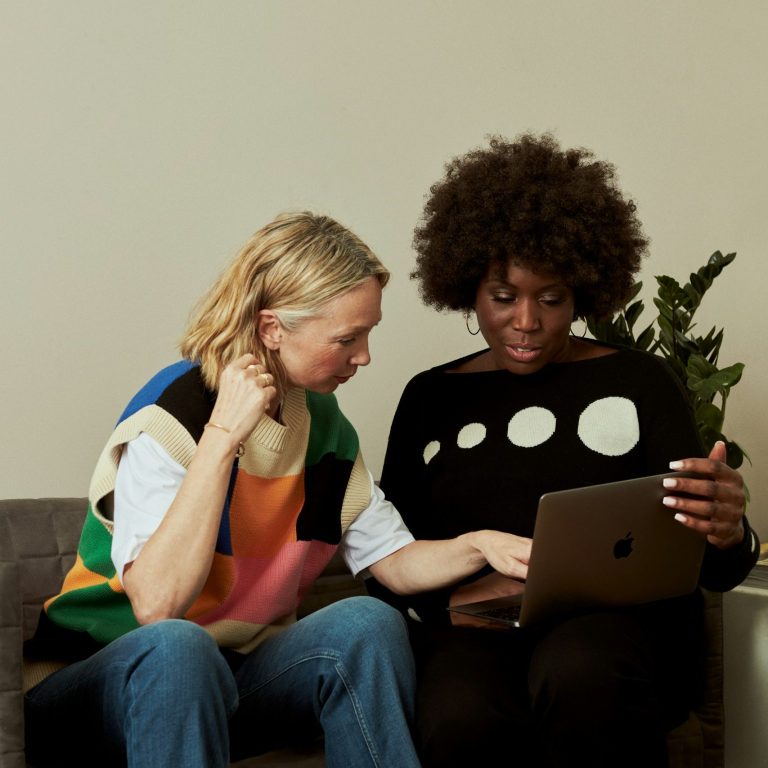 Trust and Guidance Two women sitting in a modern office lounge having a conversation, one holding a notebook and the other holding a coffee mug.