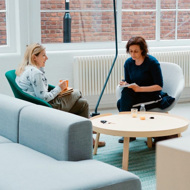 Trust and Guidance Two women sitting in a modern office lounge having a conversation, one holding a notebook and the other holding a coffee mug.