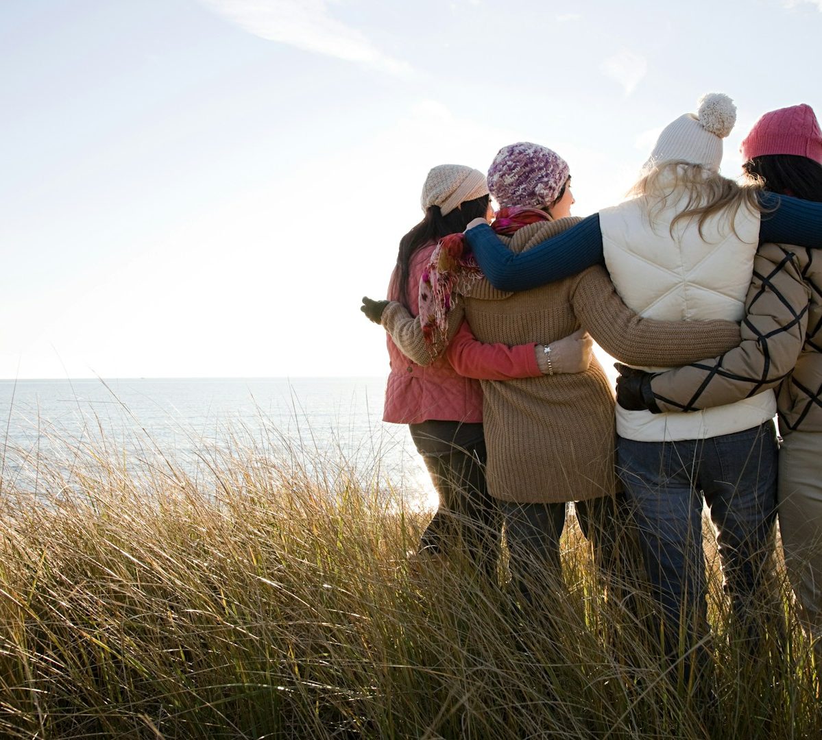 Referral Only Picture Four people standing arm-in-arm, facing the ocean on a sunny day, dressed in winter clothing and hats, surrounded by tall beach grass.